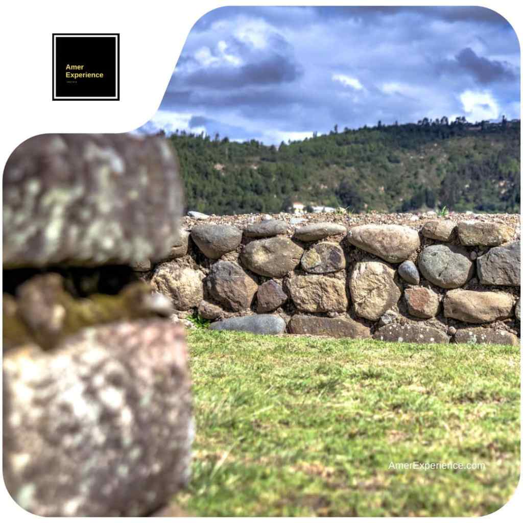 Weathered stone walls at an archaeological site in Ecuador, with grass and wooded hills beneath a cloudy sky.