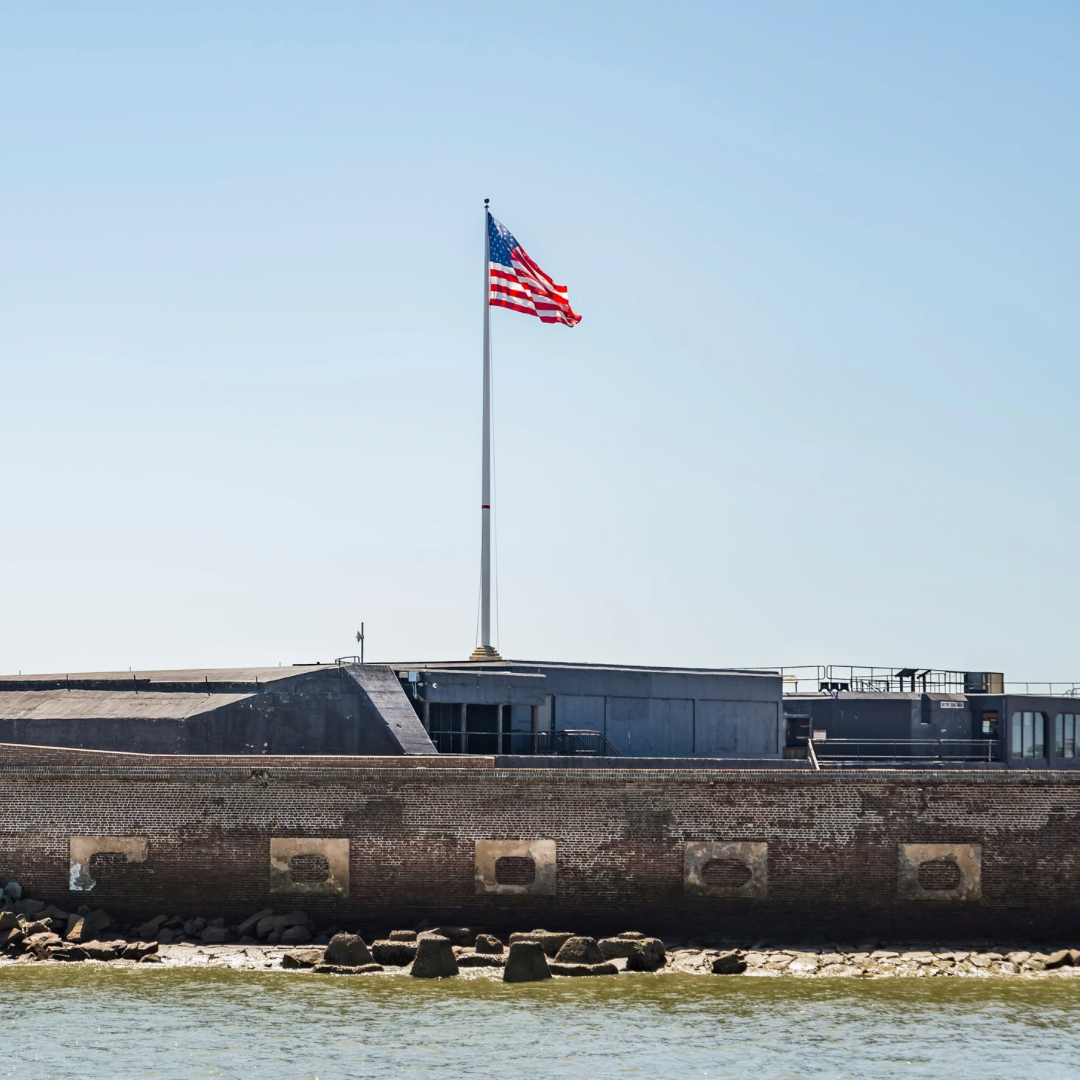 A view from the water of the stone walls of Fort Sumter National Monument with a large American flag flying against a clear sky in Charleston, South Carolina.